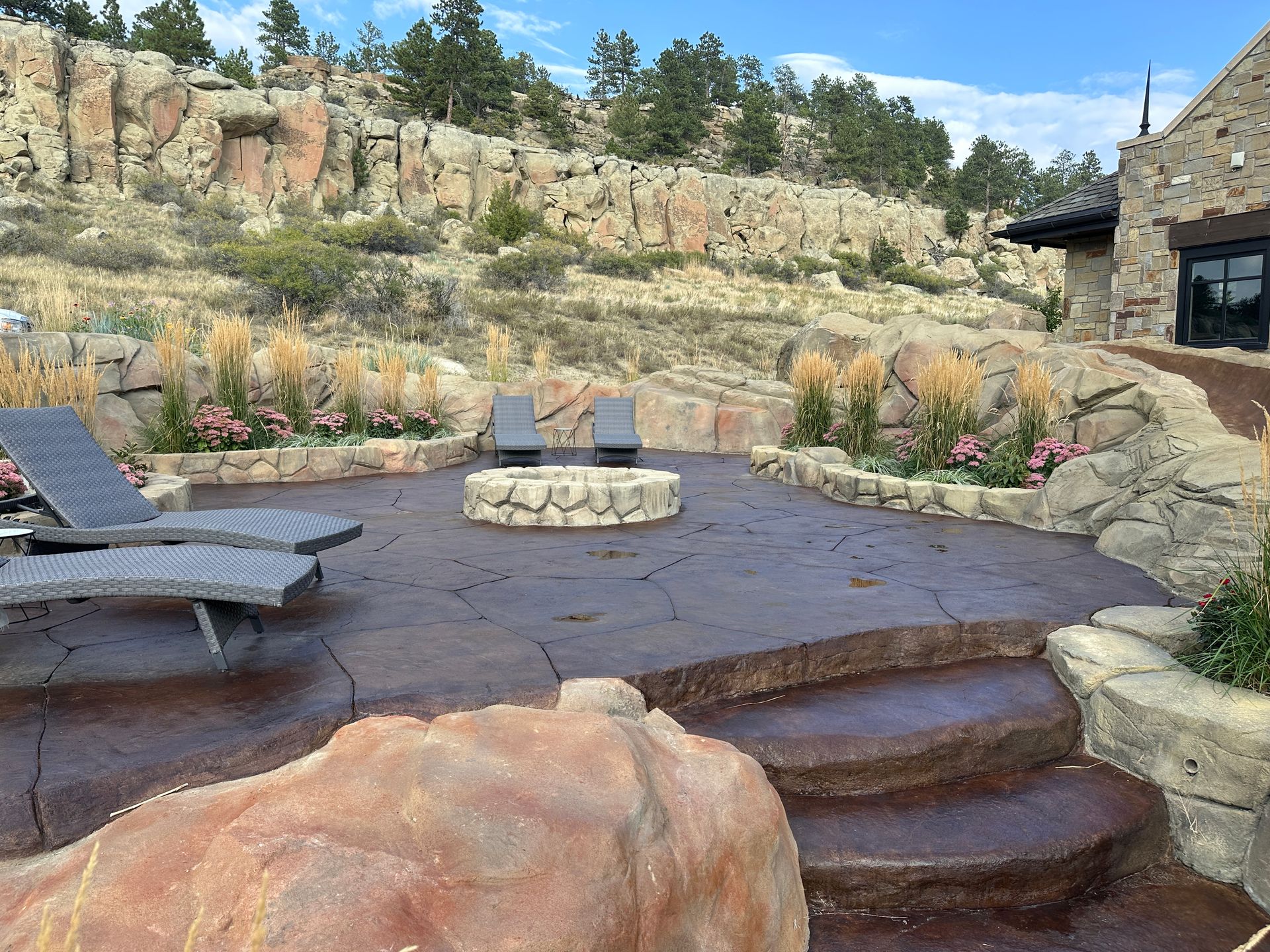 Stone patio with fire pit, lounge chairs, steps, and rock formations in a natural setting.