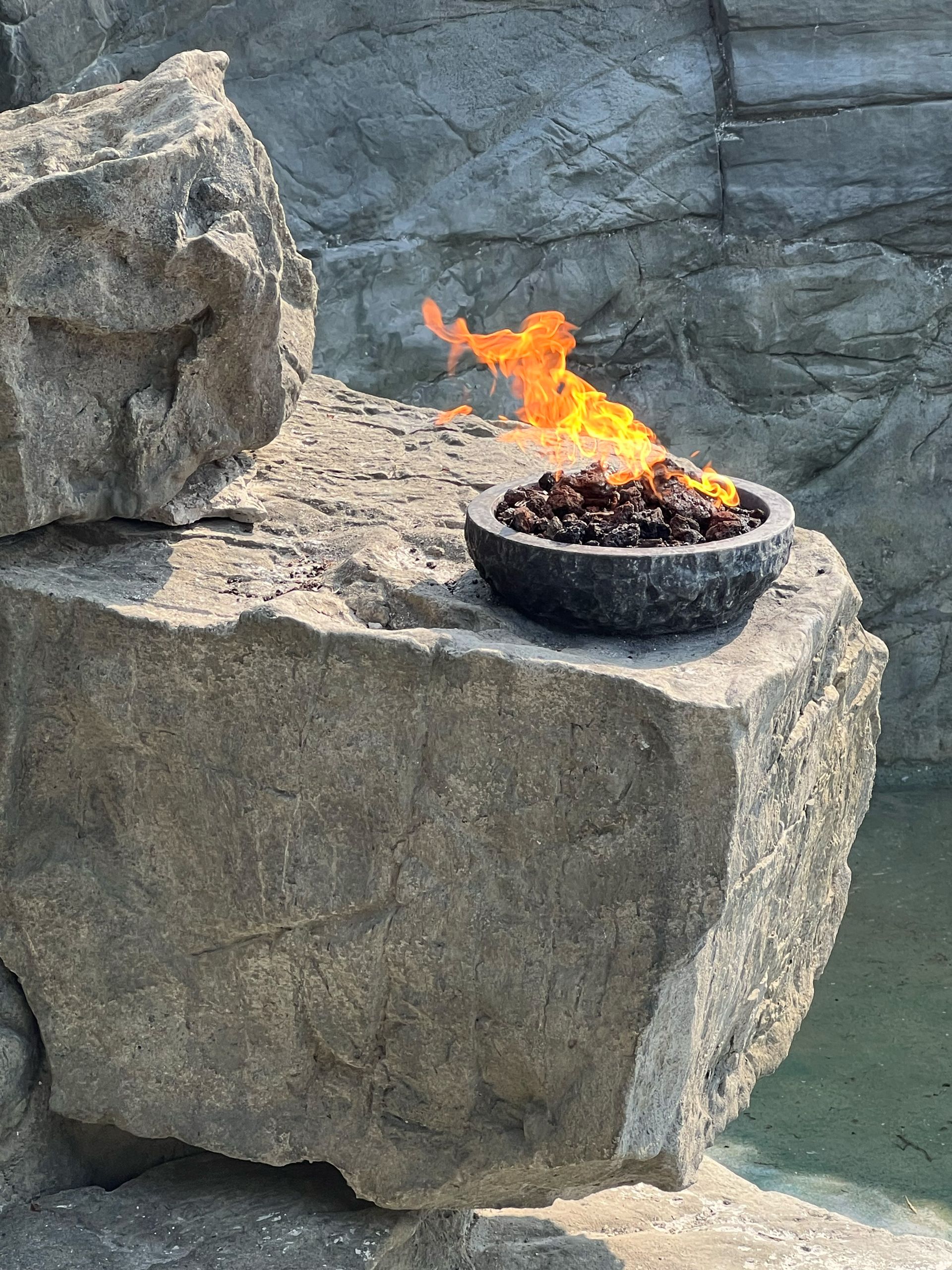Fire bowl with flames sits atop a large rock formation near water.