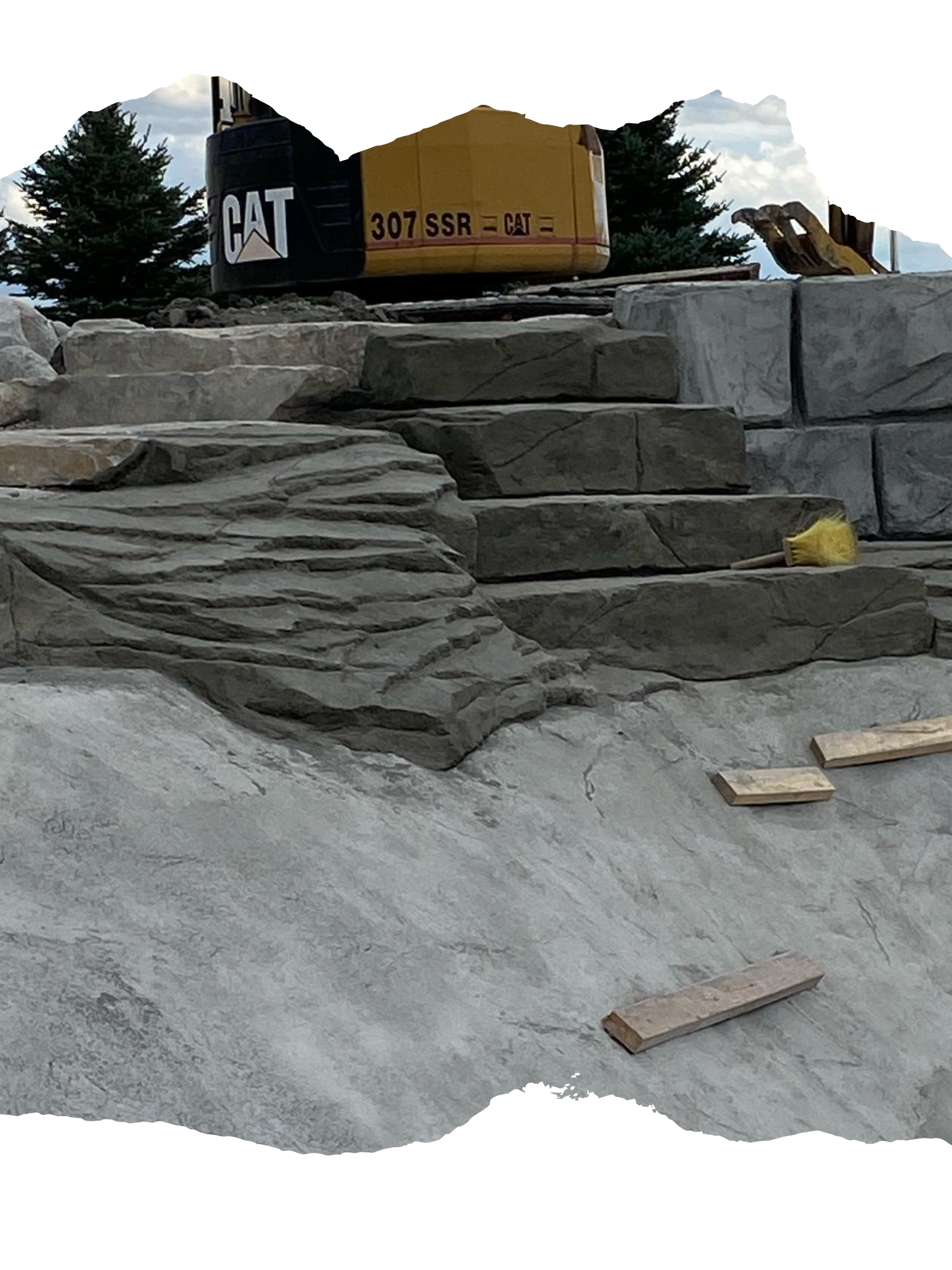 Construction site with tiered stone structure; Caterpillar excavator in background.