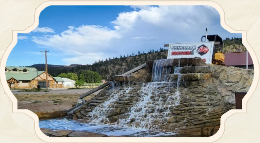 Waterfall feature with sign for a business, buildings, and mountains in the background under a blue sky.