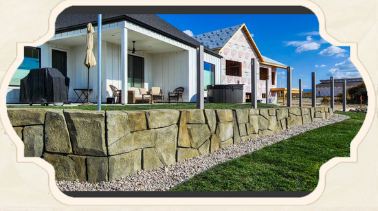 Stone retaining wall in front of white houses with black roofs, blue sky, and green grass.