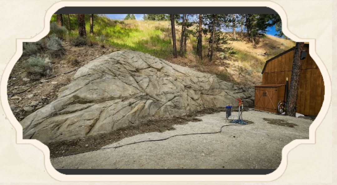 Large rock beside a wooden structure and gravel. Trees and a hill in the background.