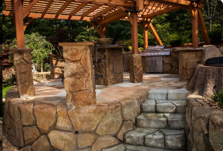 Stone patio with pergola, built-in grill, and steps. Surrounded by stone walls and landscaping.