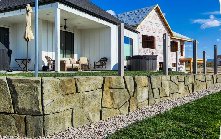 Stone retaining wall in front of white houses with black roofs, blue sky, and green grass.