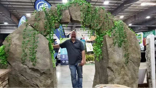 Man standing in front of large stone archway covered in greenery at a trade show.