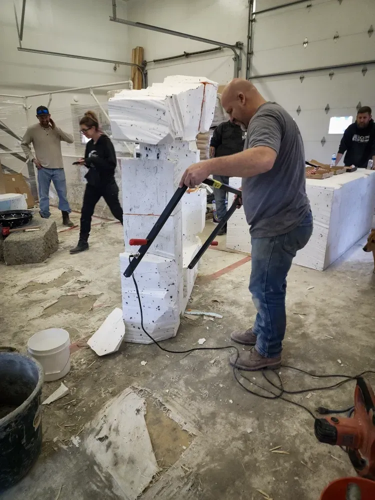 Man using tool to shape a large styrofoam structure. People in a workshop.
