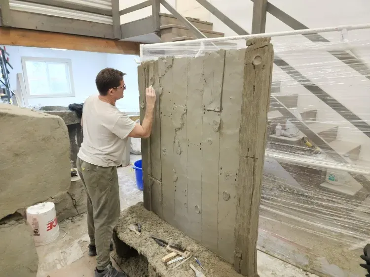Person sculpting a large, textured wall in a studio with tools and unfinished structures.