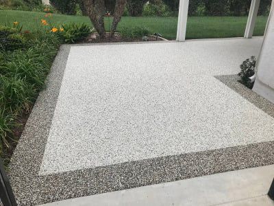 Patio with a speckled white and gray surface, bordered by a darker gray strip. Green grass and plants are visible.