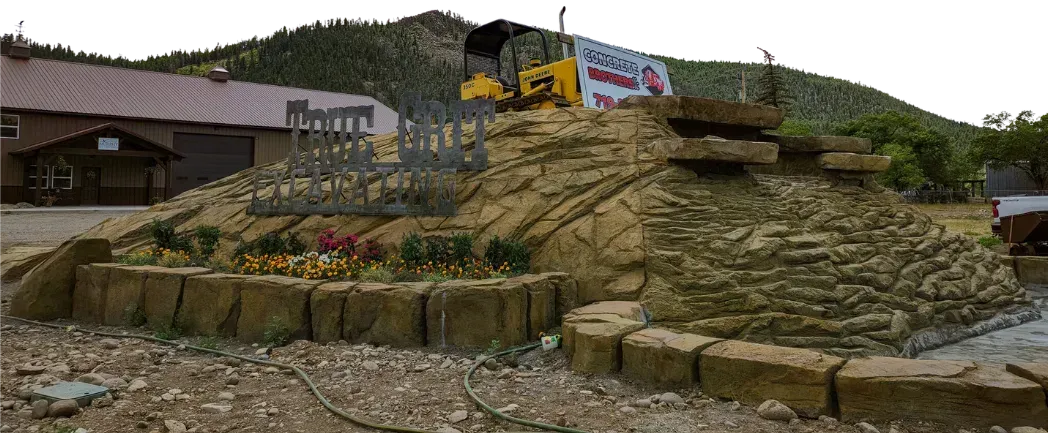 A yellow construction vehicle atop a tiered, stone-like display in front of a building and a hillside.