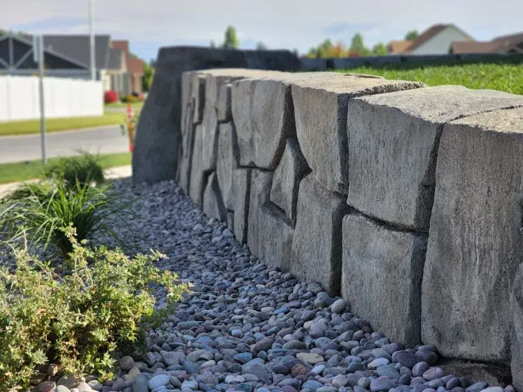 Stone retaining wall with a gravel bed, in front of a green lawn and houses.