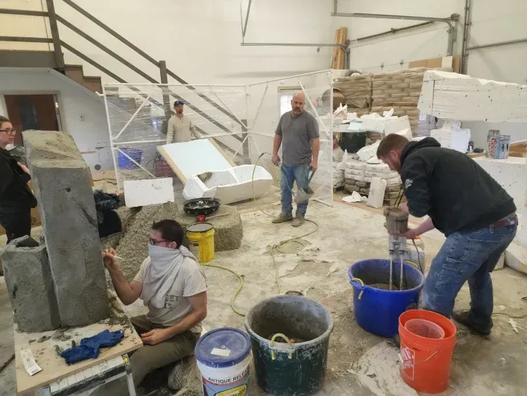 People working in a workshop. One mixes cement, another works on a concrete form, others observe.