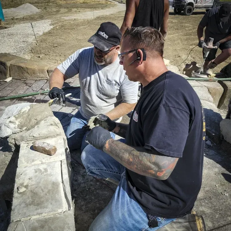 Two men working with stone outdoors, one wearing a hat and the other with tattoos, both wearing gloves.