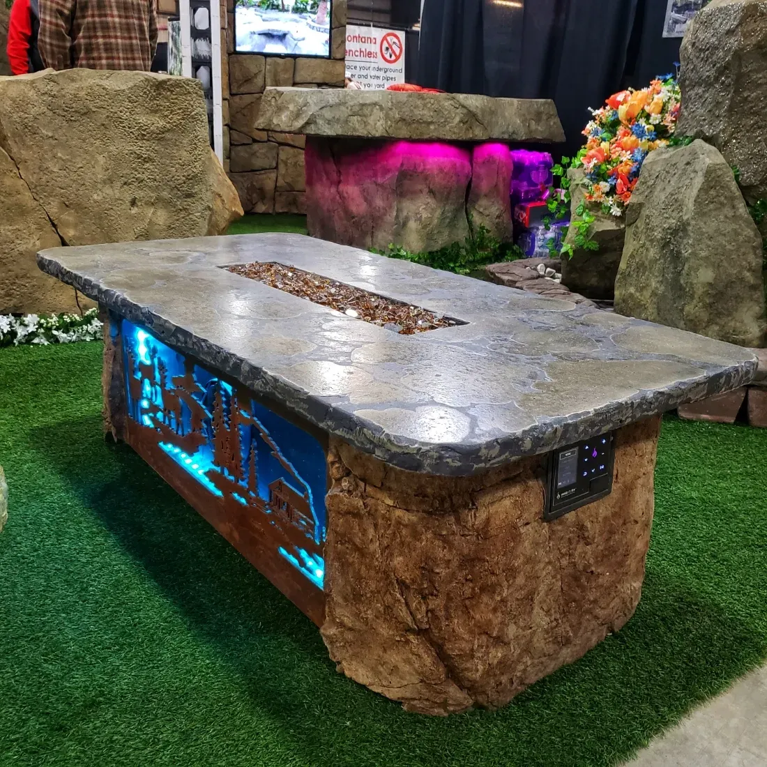 Stone fire pit table with a blue lighted facade, on green turf, at a trade show.