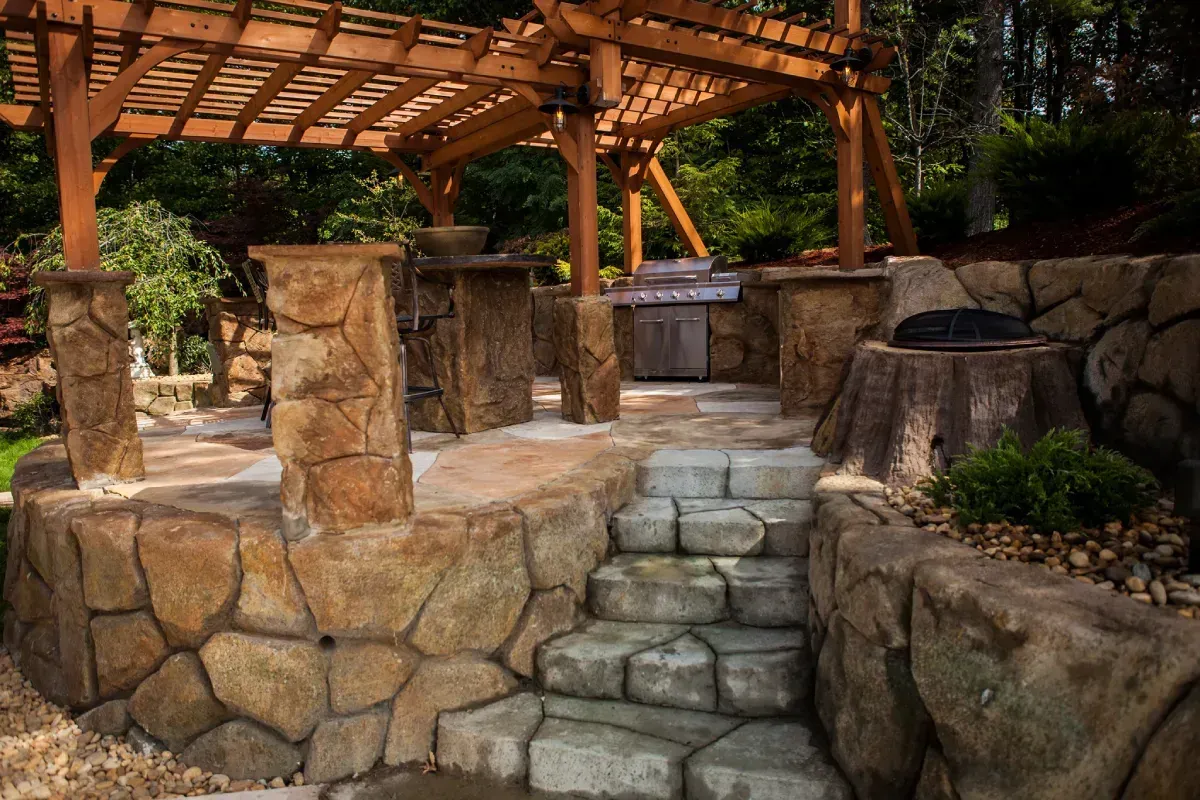 Stone patio with pergola and outdoor kitchen, featuring steps and rock walls.