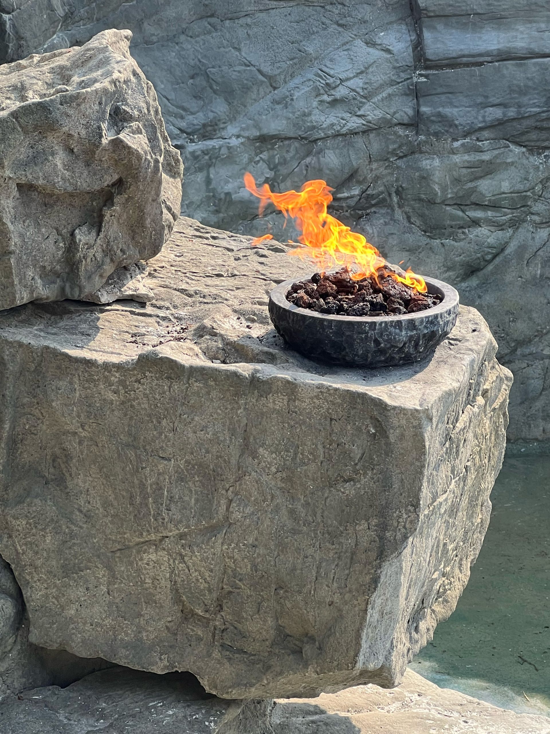 Fire burning in a dark stone bowl on a large gray rock with a rocky backdrop.