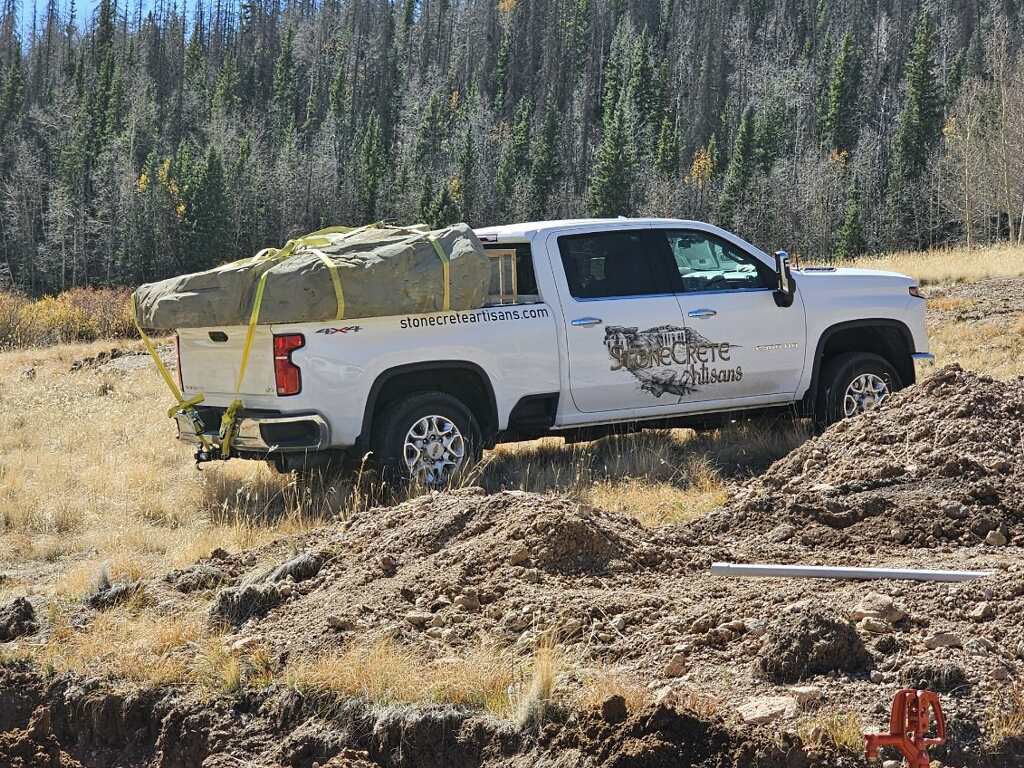 White pickup truck carrying a large rock on a hillside. The truck is off-road and surrounded by dirt.