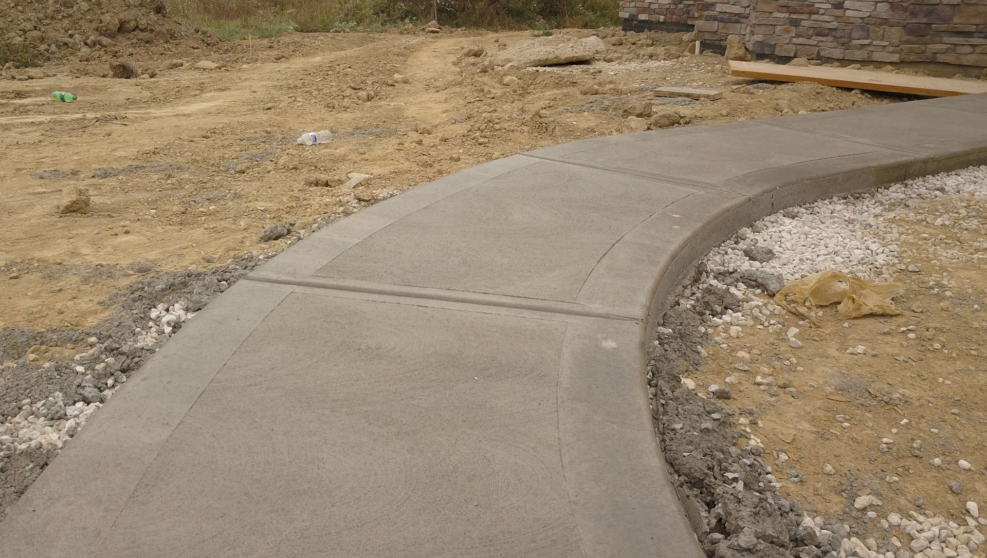 Curved concrete sidewalk under construction, surrounded by dirt and gravel.