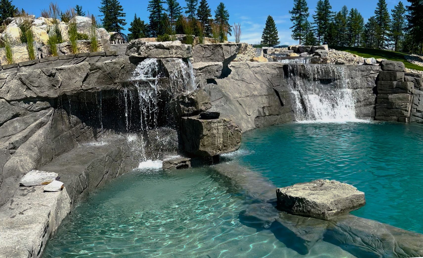 Stone waterfall cascading into a turquoise pool. Trees and sky in the background.