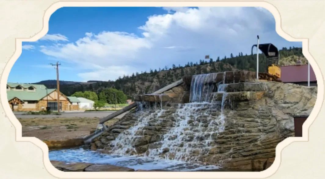 Waterfall feature with sign for a business, buildings, and mountains in the background under a blue sky.