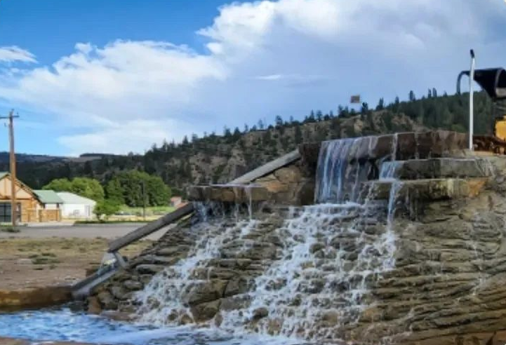 Waterfall feature with sign for a business, buildings, and mountains in the background under a blue sky.