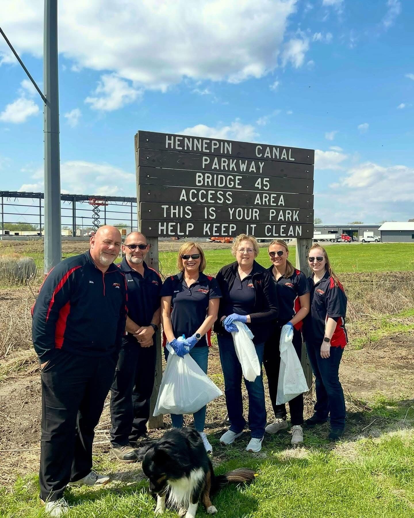 A group of people wearing black and red shirts with trash bags, cleaning a park area, under a sign that reads 