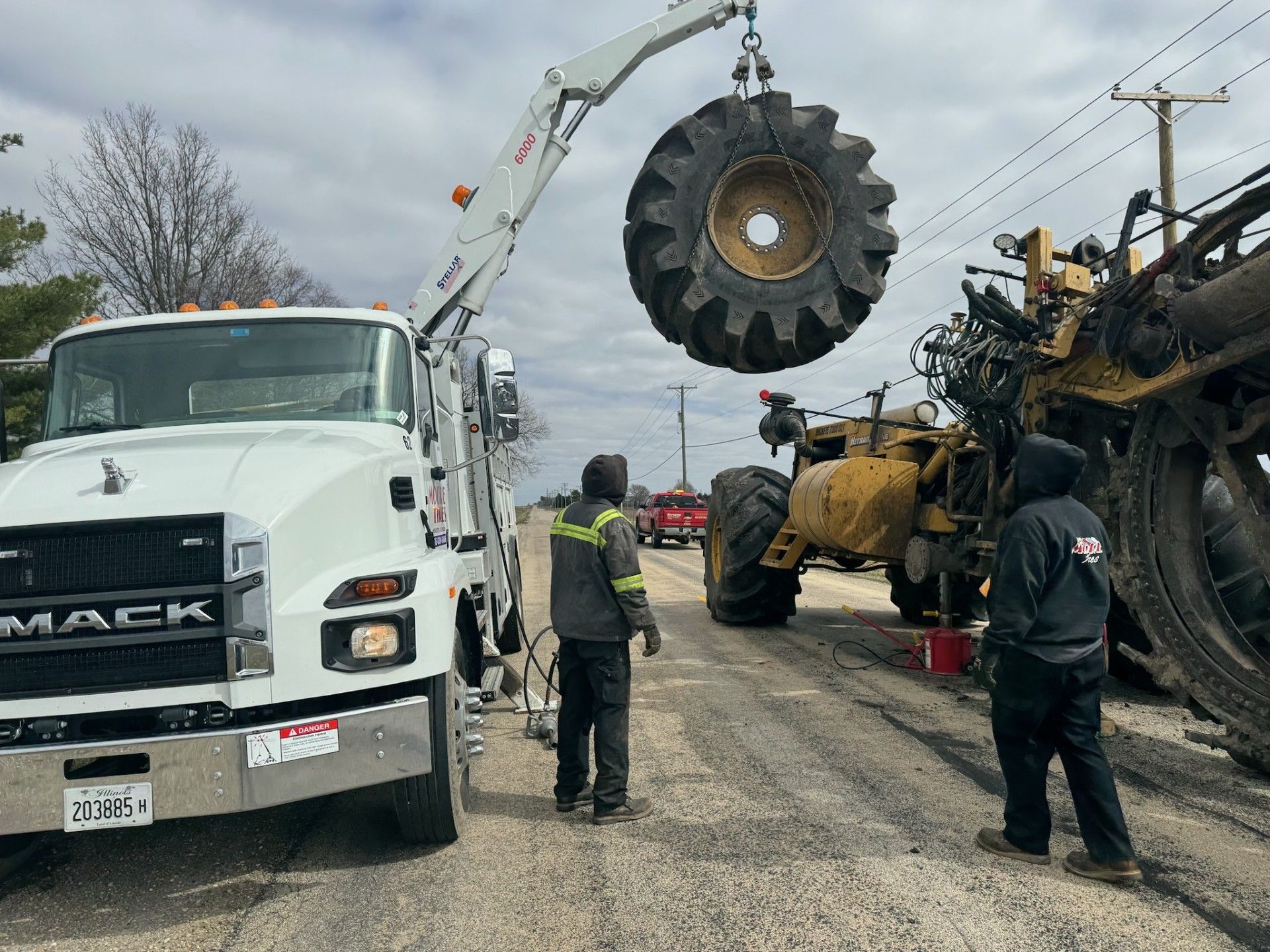 Tow truck lifting a large tractor tire on a road. Two workers stand near the tractor. Cloudy sky.