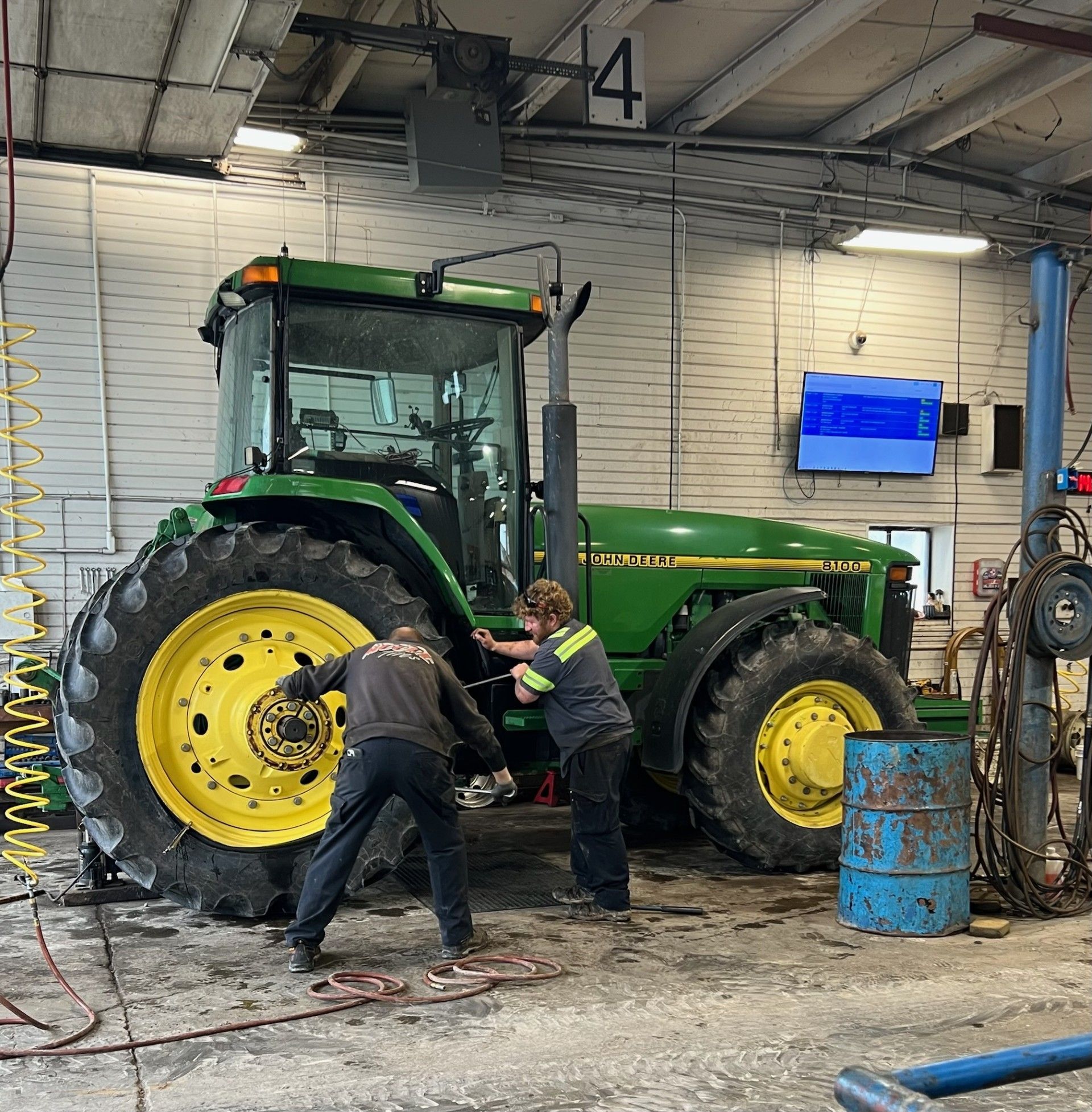 Two people work on a John Deere tractor in a garage. Green tractor, yellow wheels, blue barrels.