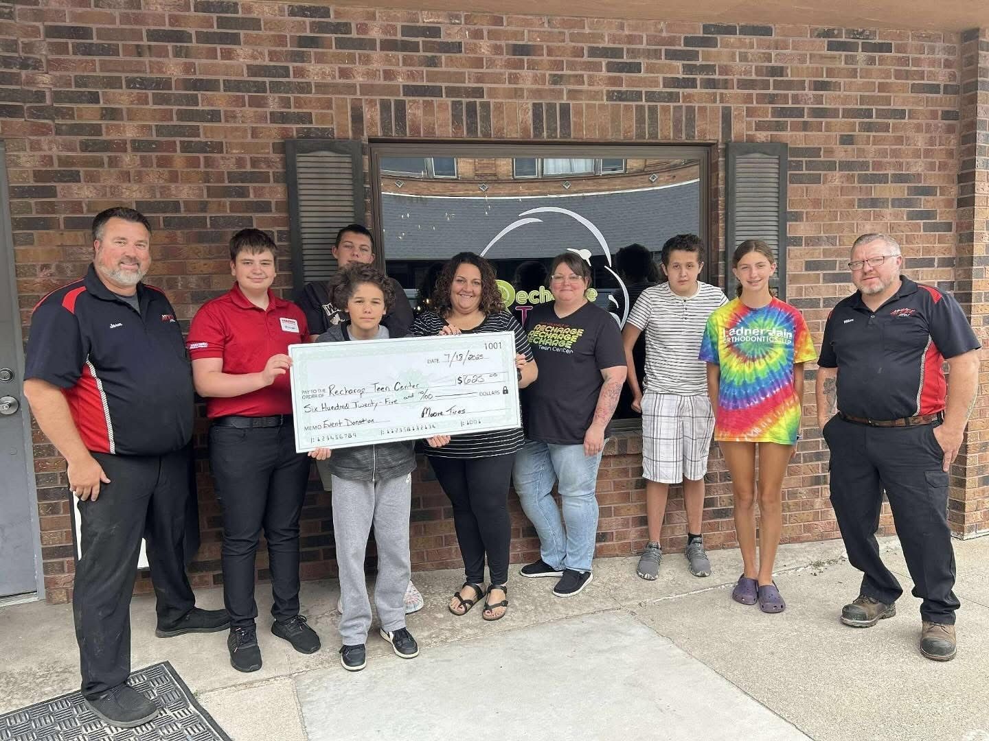 Group of people holding a large check outside a brick building.