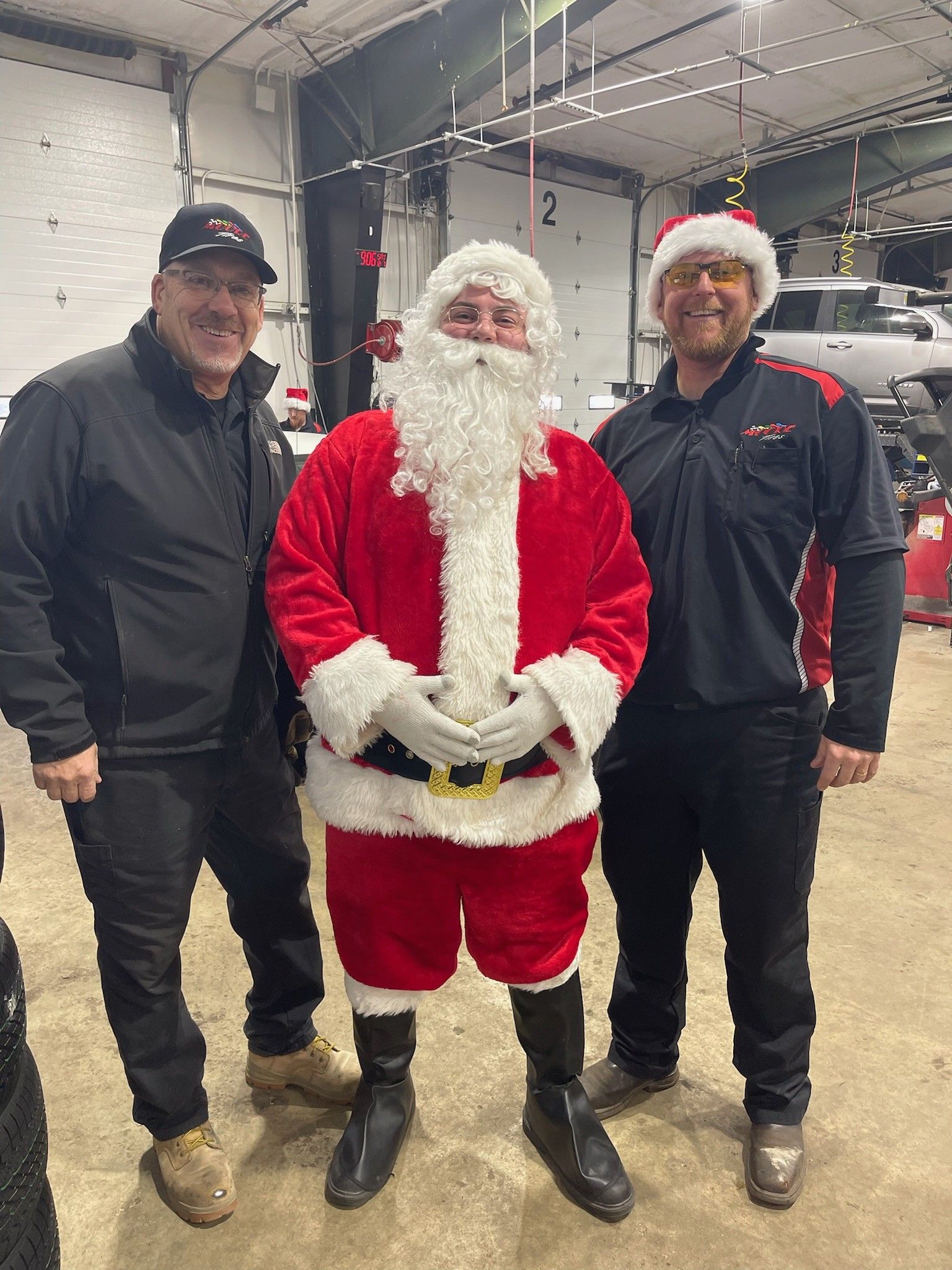 Three people posing; Santa in a red suit stands between two men. All are smiling in an industrial setting.