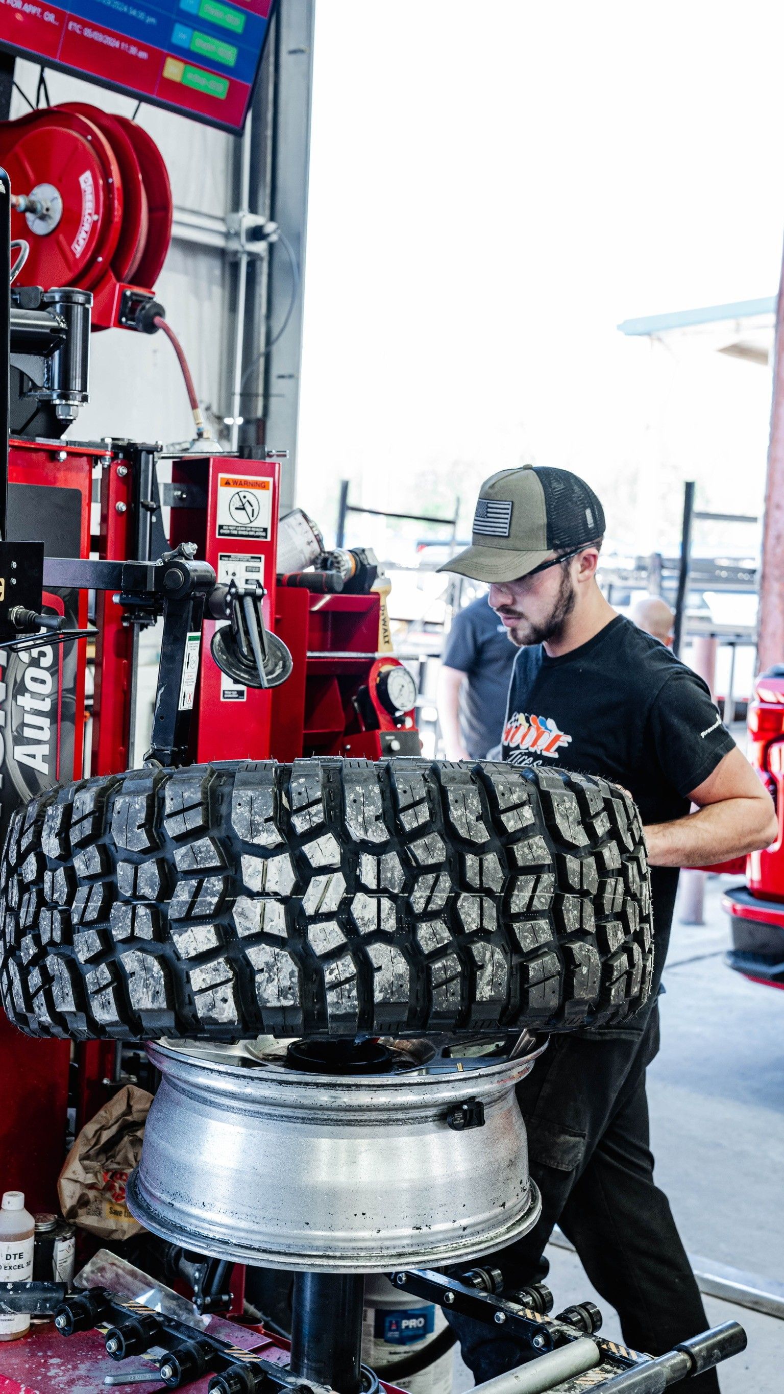 Man changing a large tire at a garage. He wears a cap and works at a tire machine. Red equipment and a bright window are in the background.