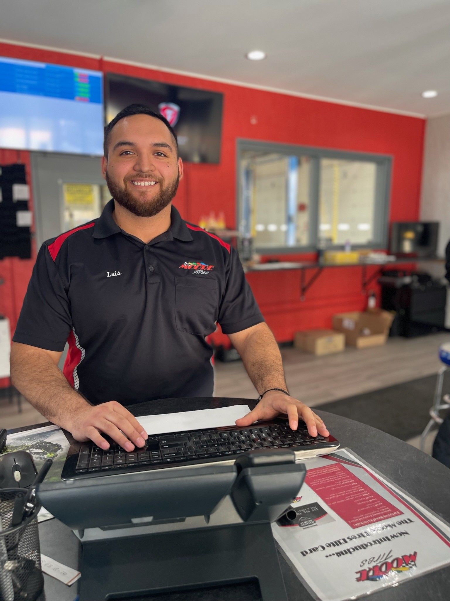 Man in auto shop uniform at a counter, smiling while typing on a keyboard. Red and black decor in the background.