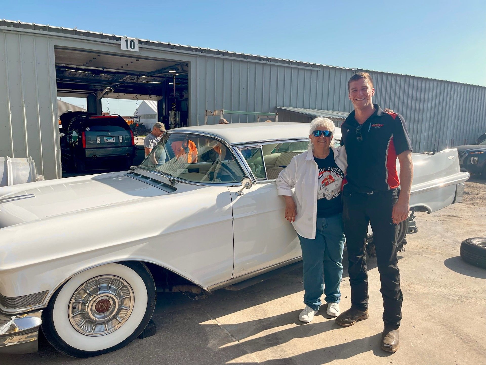 Woman and mechanic stand beside a classic white car in front of a garage.