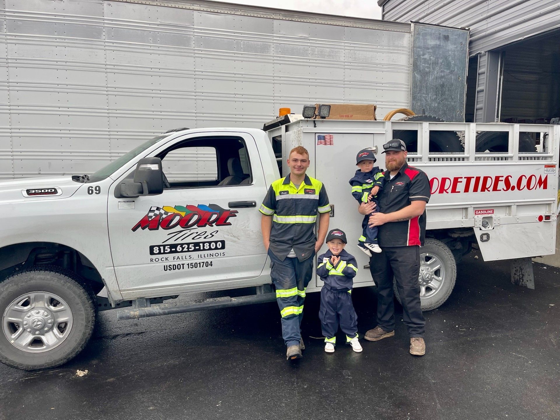 Two men and two young children pose beside a white truck with “Moore Tires” logo.