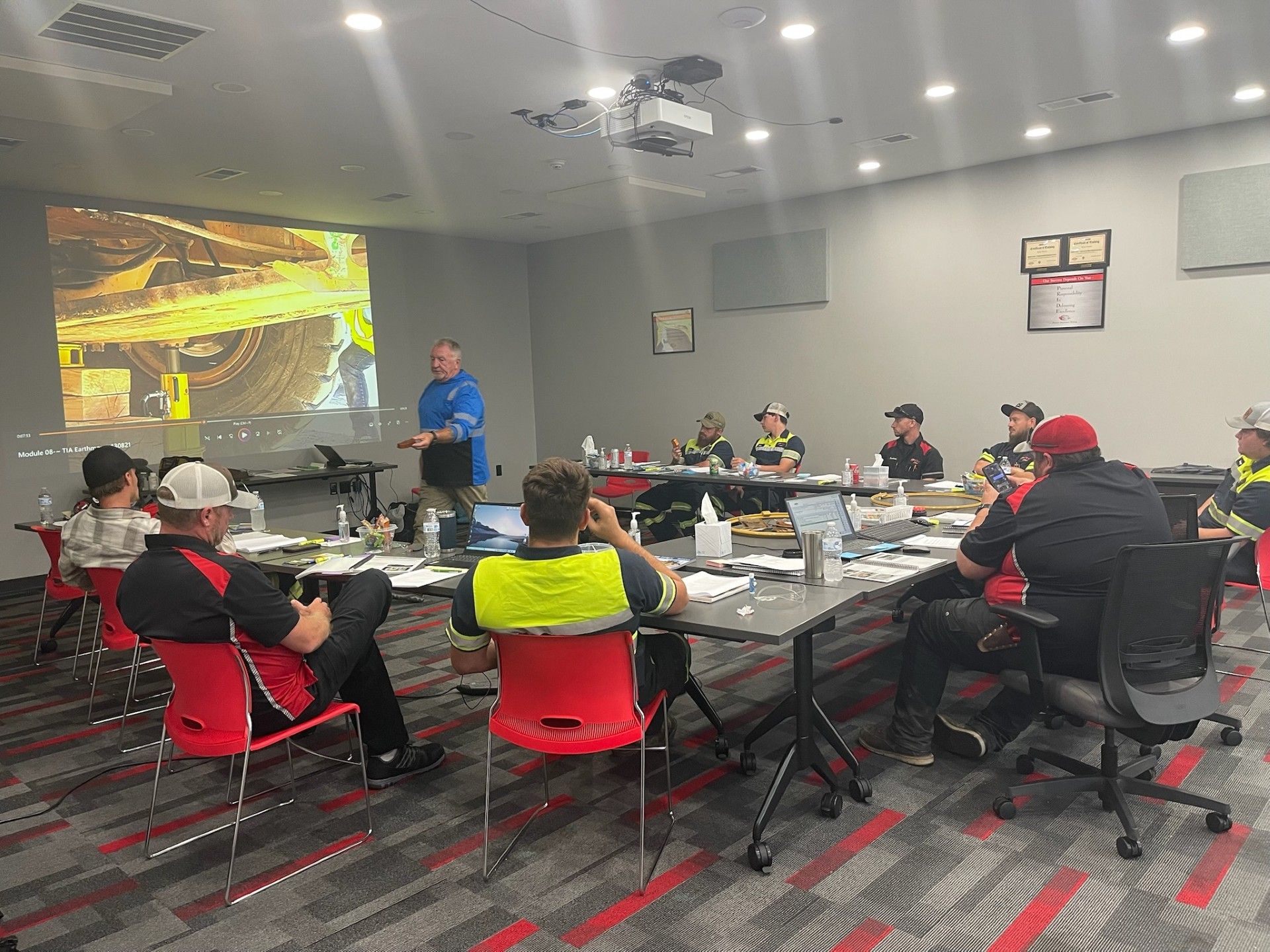 A man presents to a group seated around a table, a projector displaying a construction site.