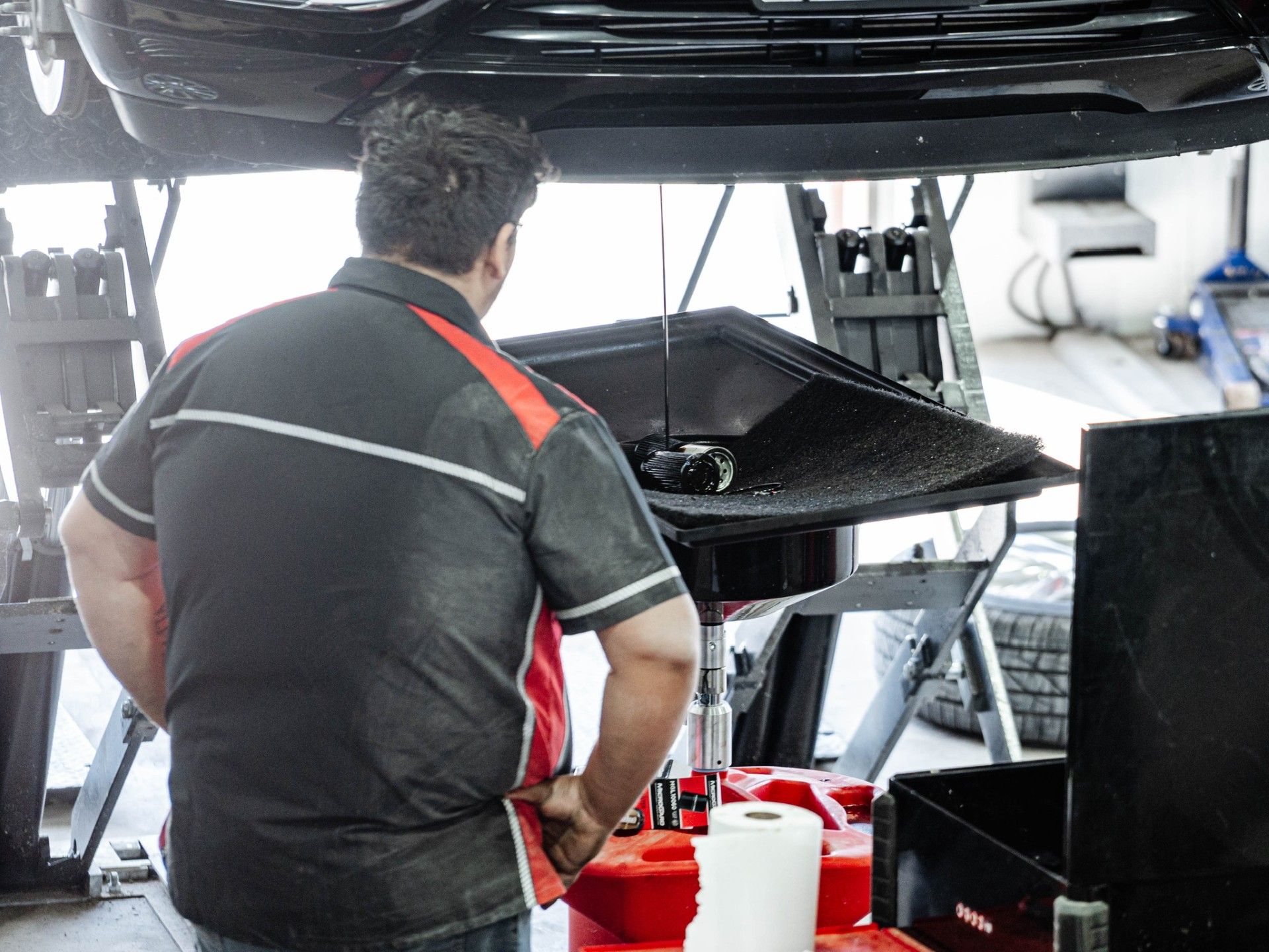 Mechanic changing oil under a car, wearing a black and red polo shirt.