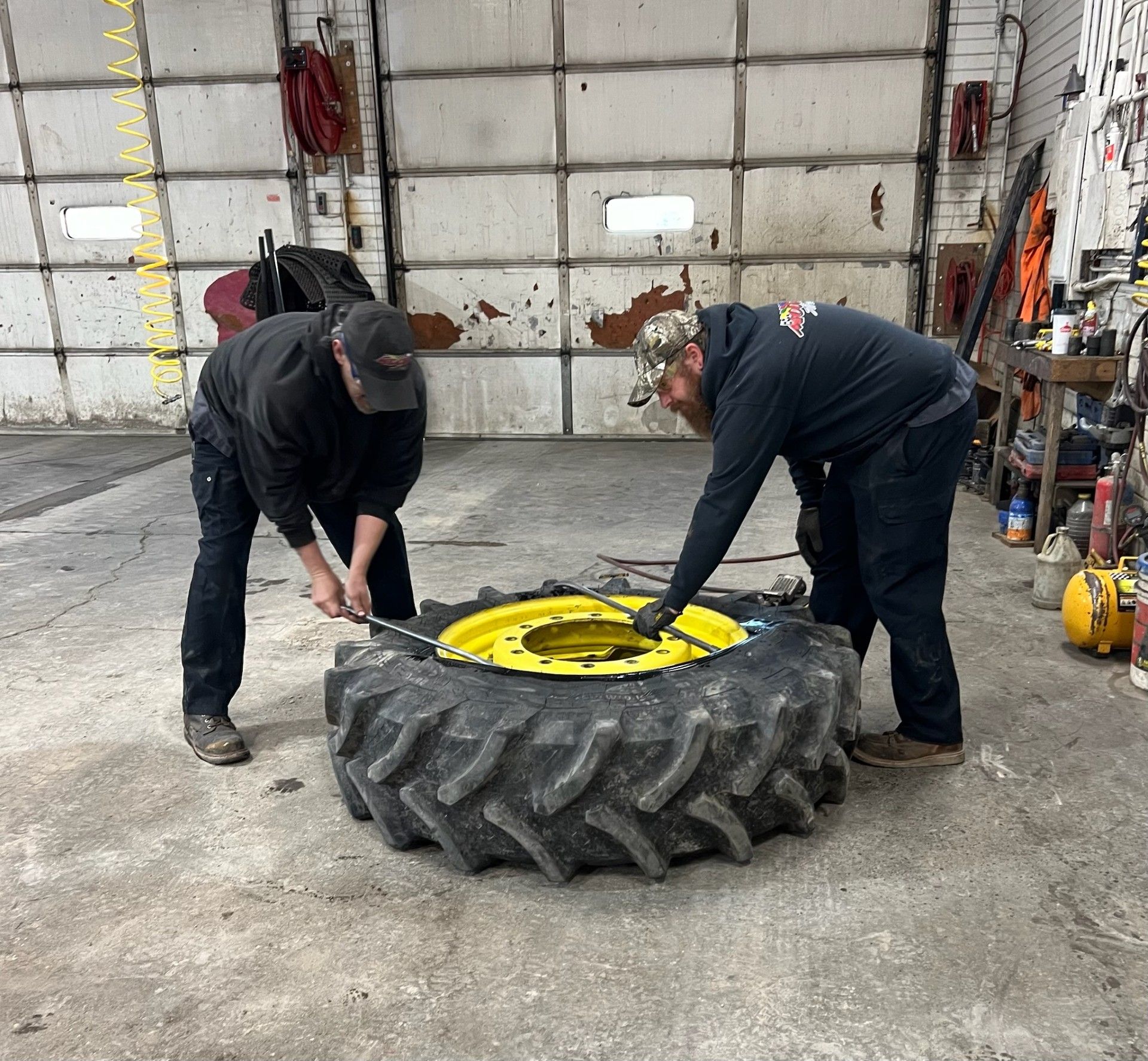 Two people working on a large tractor tire in a garage, one using a tool.