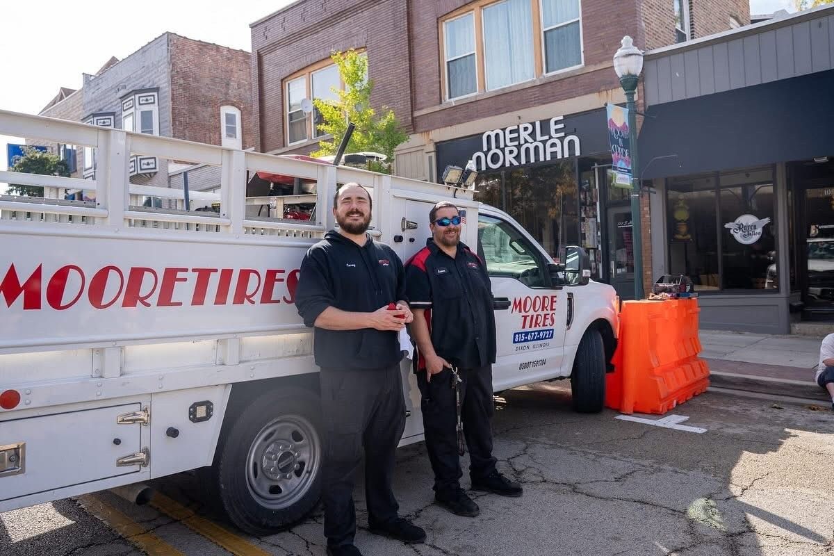 Two men stand by a Moore Tires truck on a street. One smiles. A Merle Norman sign is visible.
