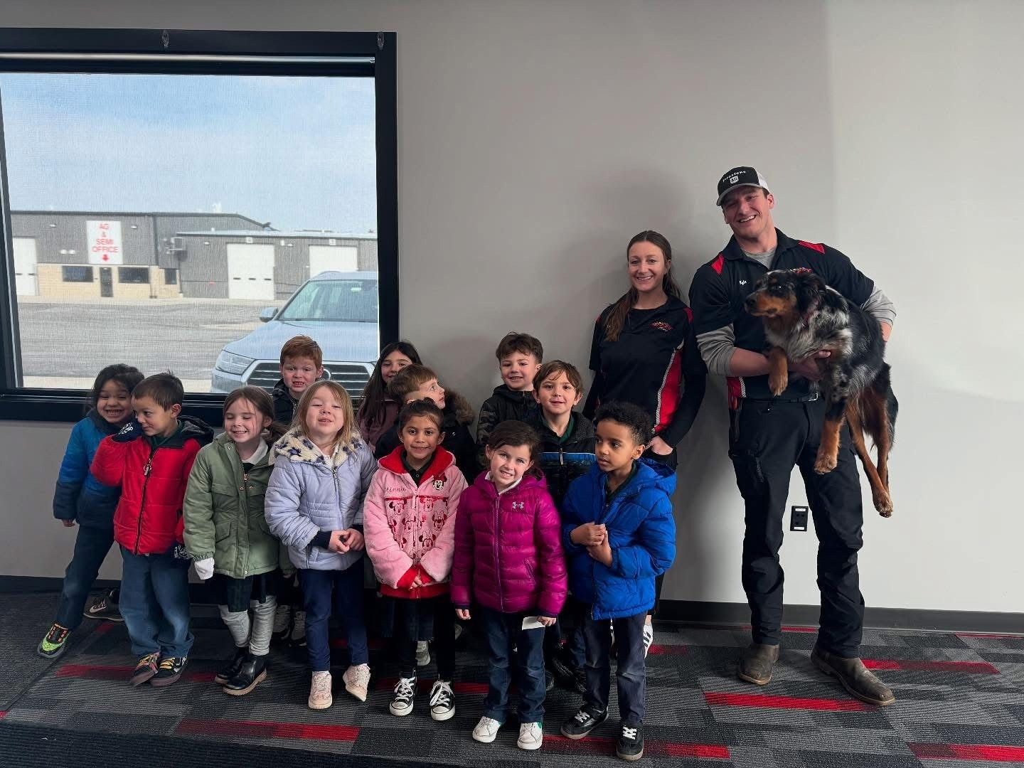 Group of children with two adults, one holding a dog, near a window, inside a building.