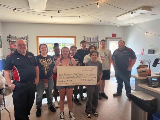 Group holding a large check, smiling in an office. Red and white accents, fluorescent lighting.
