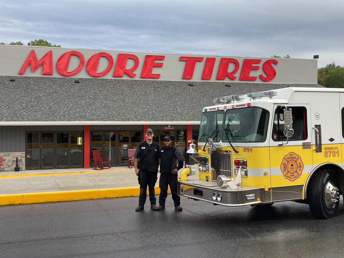 Two firefighters stand in front of a Moore Tires store next to a yellow and white fire truck.
