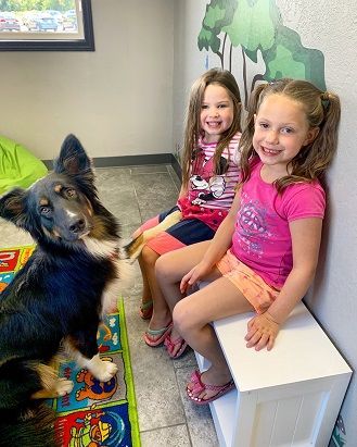 Dog with two young girls sitting on a bench in a waiting room, smiling.