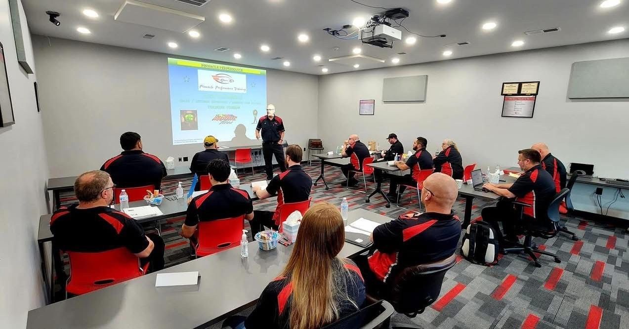A group of people in red and black uniforms attend a training session in a classroom. A presenter stands at the front.