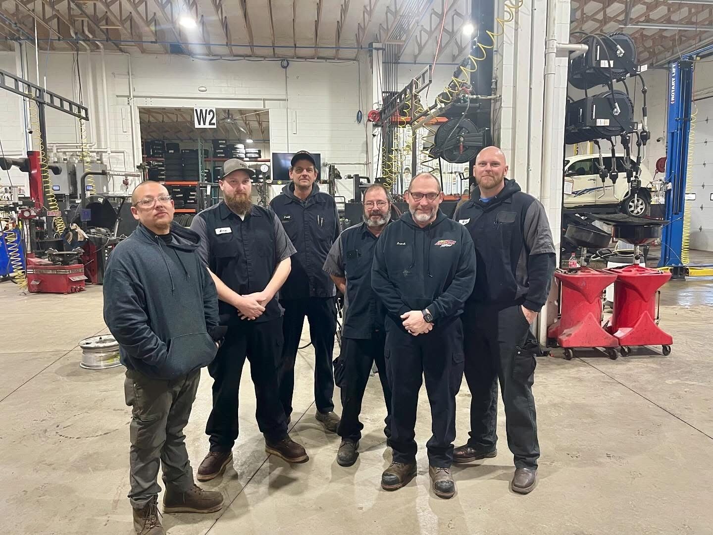 Group of seven people posing in an auto repair shop. The people are in work clothes and appear to be mechanics.