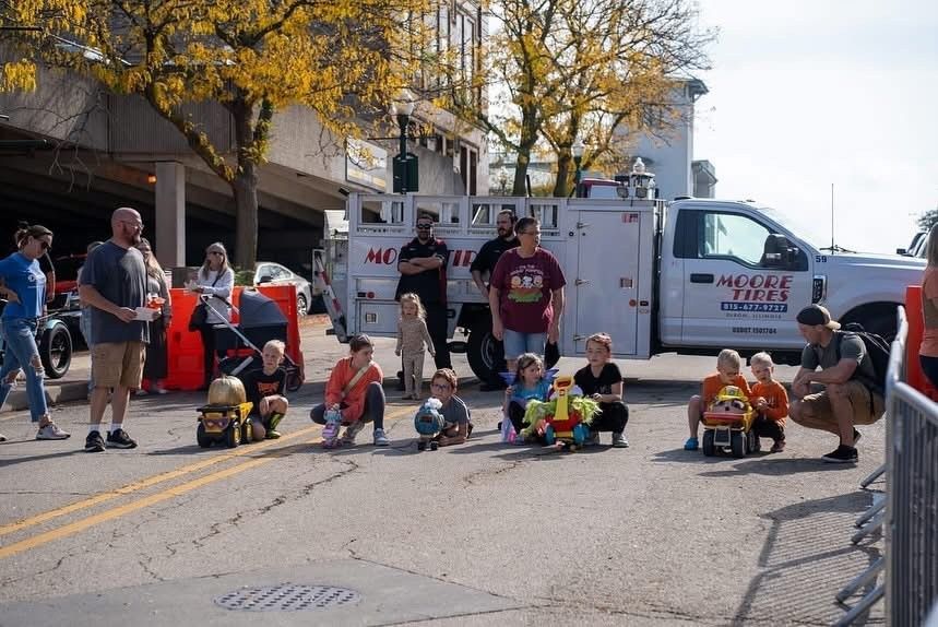 Children on toy cars line up on a street for a race, with a crowd and a utility truck in the background.