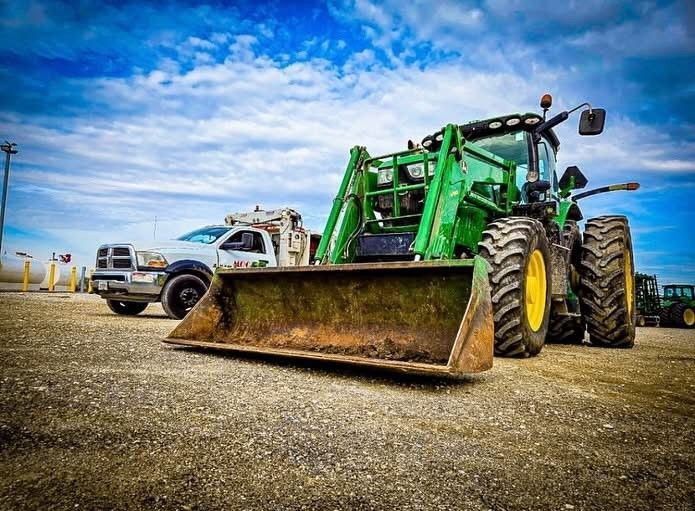 Green tractor with a front-end loader parked on gravel. A white truck sits to the left under a cloudy sky.