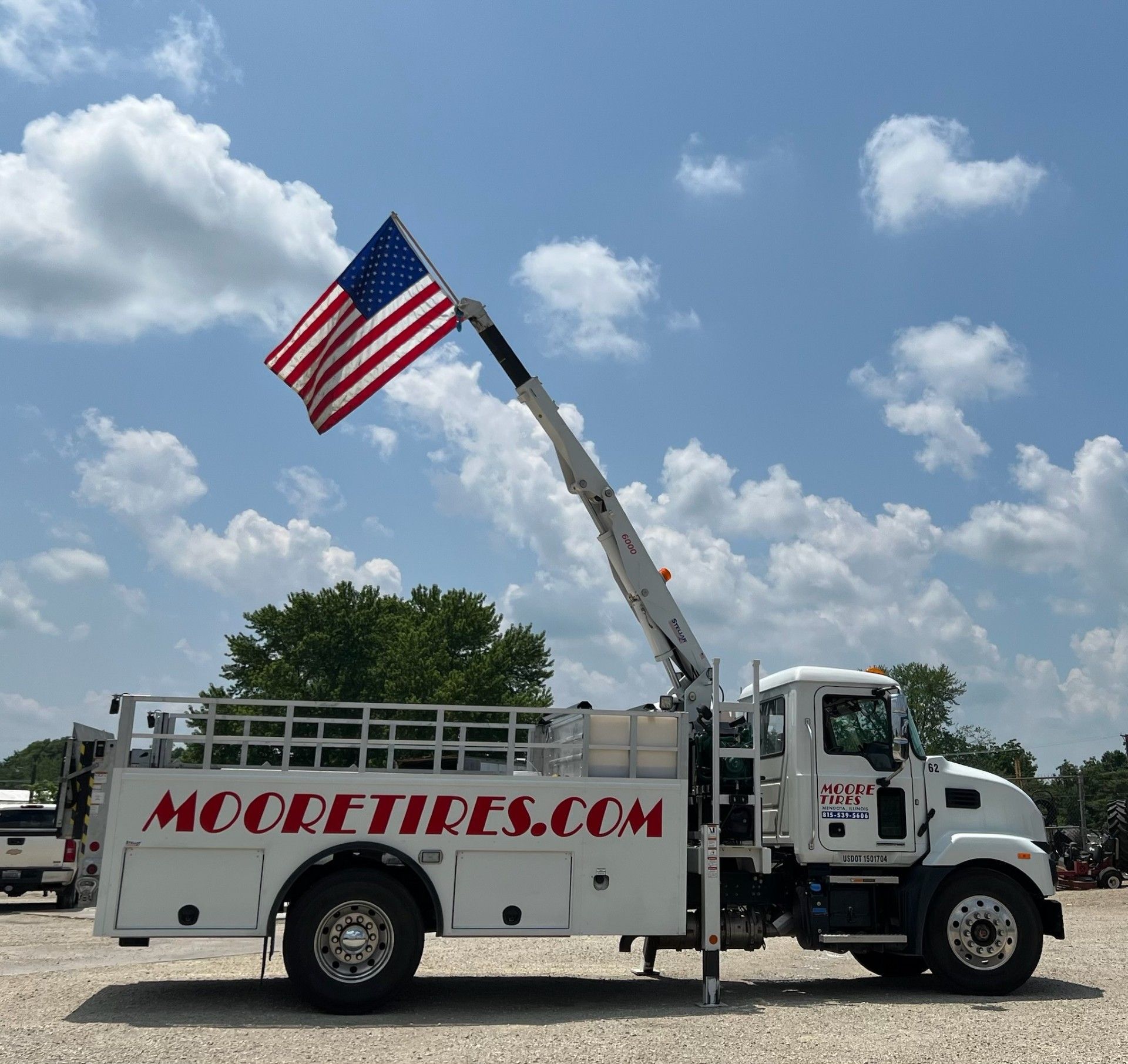 White Moore Tires truck with American flag extended by a crane against a blue sky.