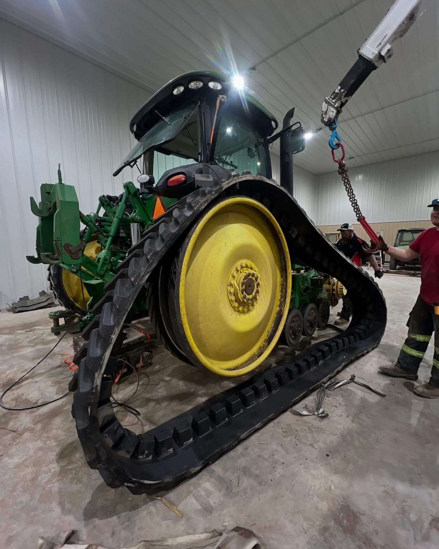 A John Deere tractor with tracks being lifted by a crane in a warehouse.