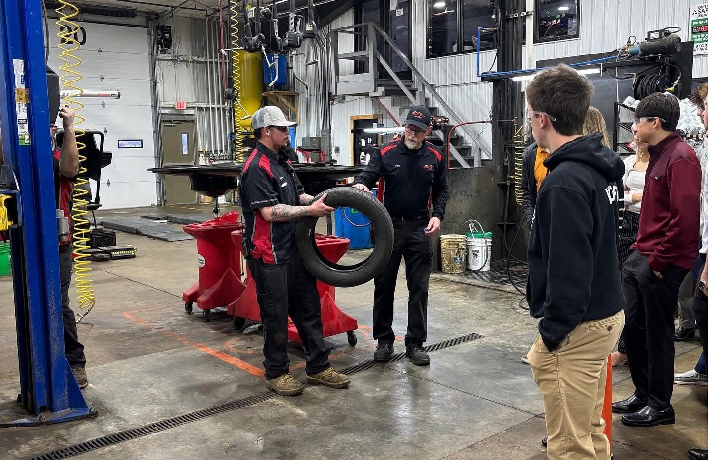 Mechanic holding a tire, instructing students in a garage.