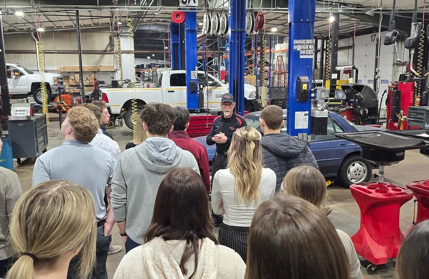 Group of students in an auto shop listening to a man speaking. Vehicles and equipment surround them.