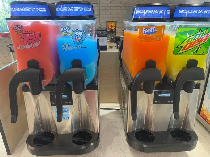 Four colorful slushy dispensers: red, blue, orange, and green. Black handles. White countertop.